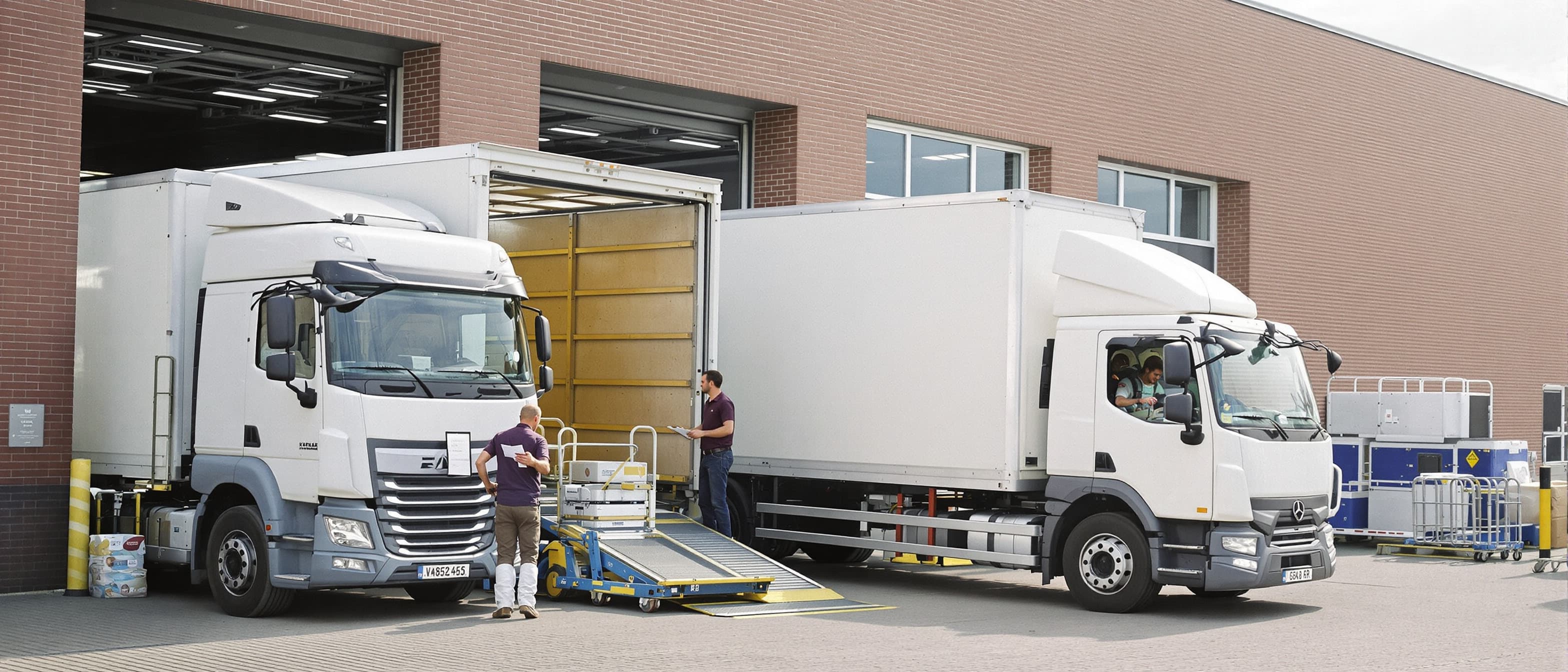 Twee bakwagens staan aan het laadperron van een Nederlands bedrijfspand, terwijl crew karren de laadklep op rijdt en een planner met een laadlijst bij de cabine staat
