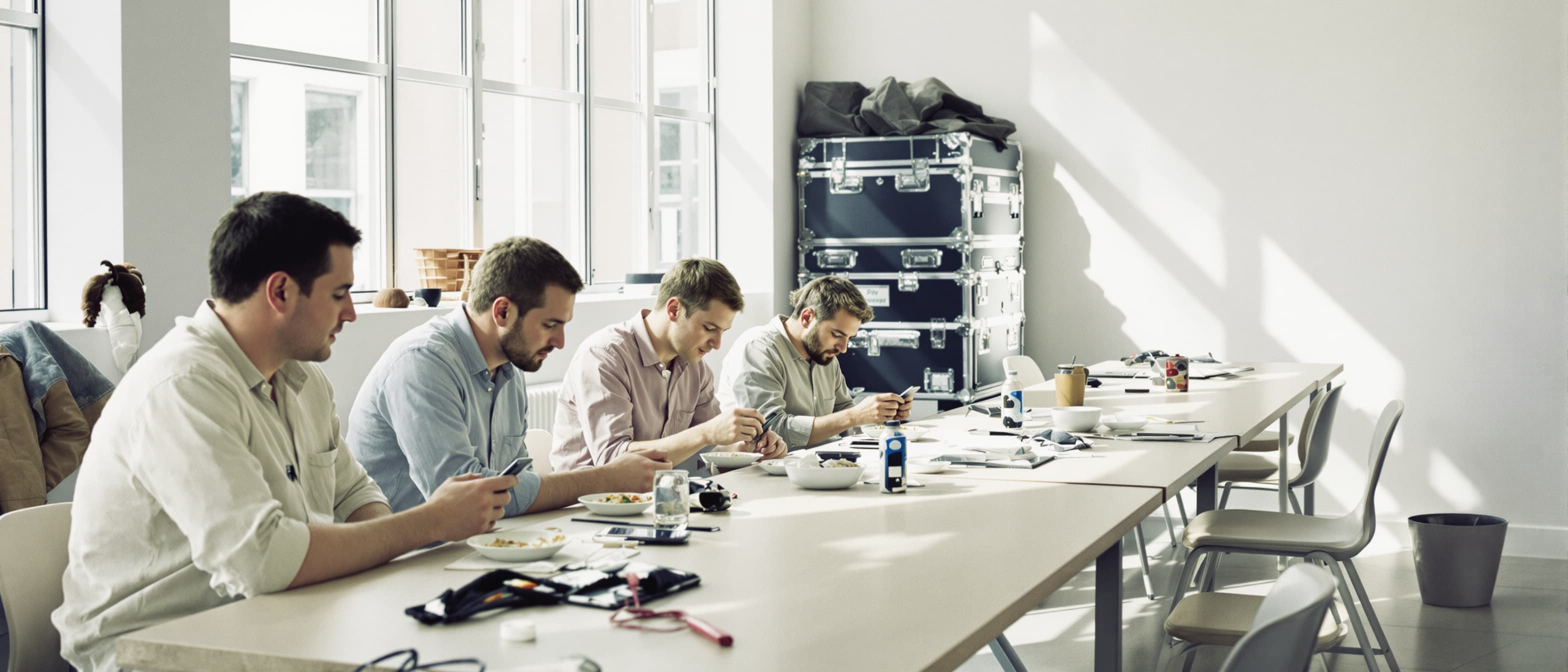 Vier AV-freelancers zitten tijdens de pauze aan een lange tafel in de crewruimte, elk verdiept in zijn eigen telefoon, met flightcases op de achtergrond